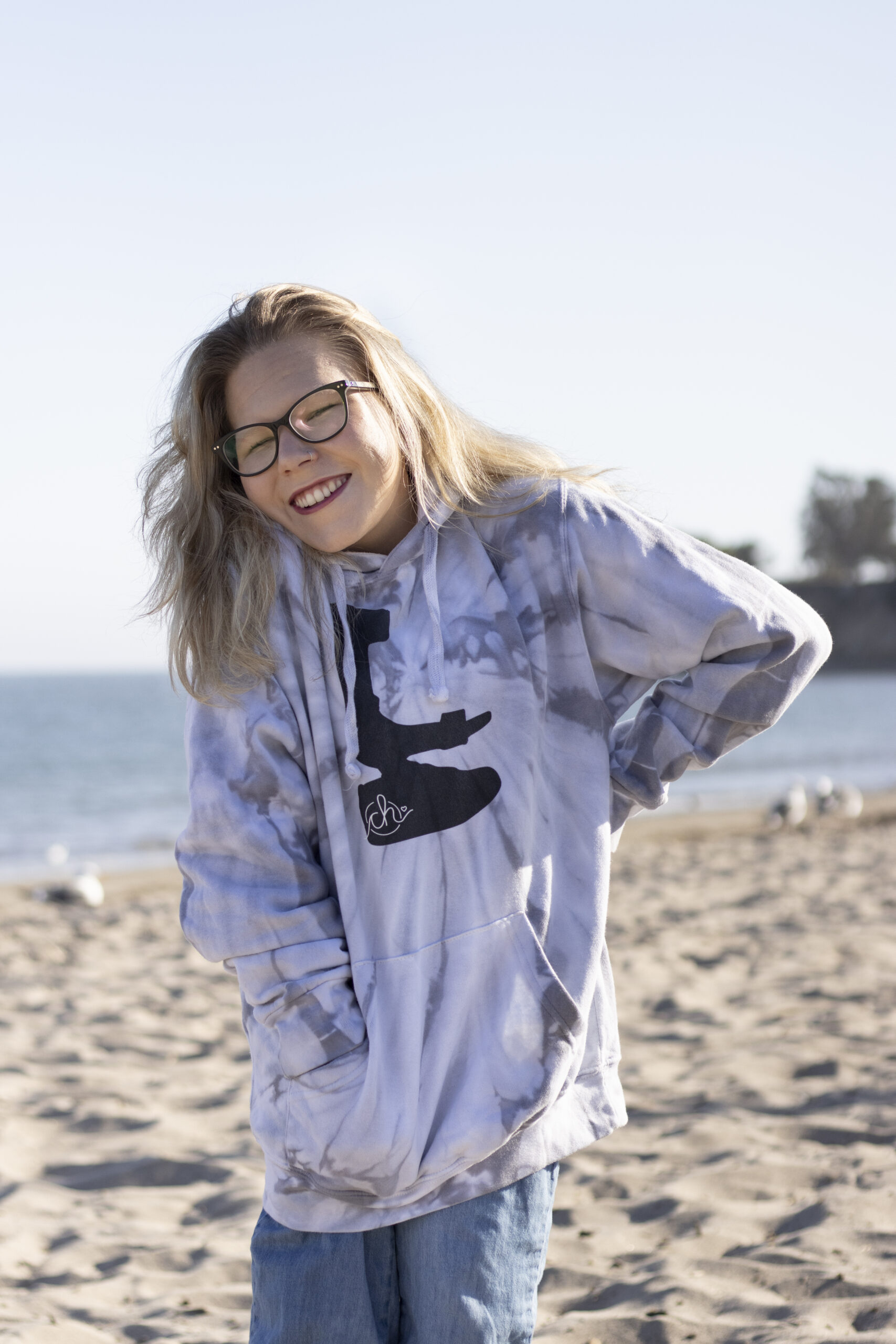 A blonde haired woman wearing a glasses and hoodie standing on the beach smiling at the camera.